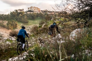 Two riders on gravel bikes pick their way along a rocky path towards a hilltop village in Jordan.