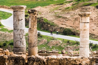 Two cyclists ride a paved road seen between ancient stone columns at a ruined site in Jordan.