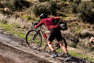 Rider in a maroon Ryzon jersey hauls a gravel bike up a steep stony bank beside a road in Jordan.