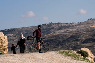 Rider stands with a bikepacking gravel bike on a Jordanian hilltop as a woman and child walk past.