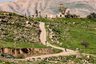 Two small figures ride a winding dirt track across green hills below bare mountains in Jordan.