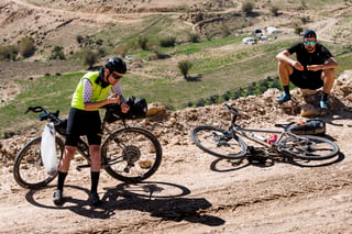 Two riders stop on a rocky ridge in Jordan, one eating a snack beside a bike, the other sitting nearby.