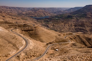 Hairpin road winds through bare hills down to a reservoir and dam on the Jordan Bike Trail route.