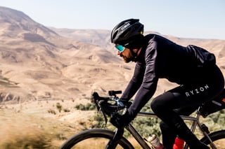Close-up of a rider in a black helmet and sunglasses pedalling a gravel bike past desert hills in Jordan.