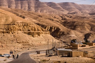 Two riders stand with bikes beside a road at a small Jordanian settlement under dry, layered hills.