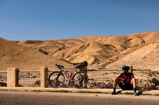 A rider sits on a kerb in the sun beside a bikepacking bike propped on a roadside rail in Jordan.