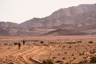 Two riders cross open desert on churned sandy tracks below dry mountains in southern Jordan.