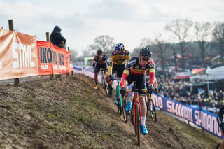 A rider in black with red and yellow bands leads a group along the banked slope at the Hoogerheide Cyclocross World Cup.
