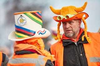 Two fans in orange hi-vis vests and novelty hats, one a rainbow-striped top hat, in the crowd at Hoogerheide.
