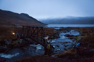Rainspotting riders with head torches at a wooden footbridge over a Highland river at dusk, a loch and snowy hills beyond.