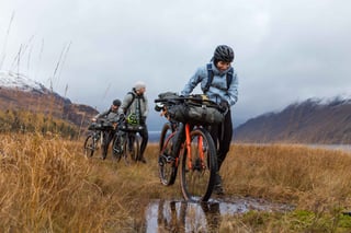Rider pushes a loaded orange bike through a muddy bog in the Highlands, with two more riders behind, during Rainspotting.