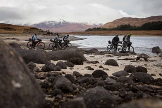 Bikepackers push their bikes across rocks and sand beside a Scottish loch in the Rainspotting film.