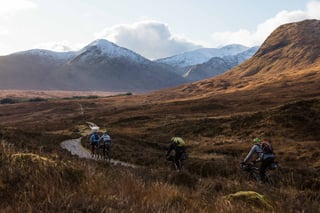 Rainspotting riders on loaded bikes follow a gravel track beneath snow-capped Highland mountains in low winter light.