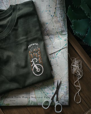 Get Wild x ADVNTR tee with embroidered bike and pine trees on a North York Moors map, needle and scissors beside it.