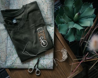 Folded Get Wild x ADVNTR tee on a North York Moors map beside a potted succulent, with scissors and loose thread.