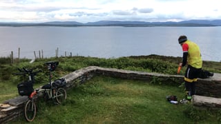 Loaded Brompton leans on a stone wall beside a wide bay as a rider pulls on gloves on the Irish coast.