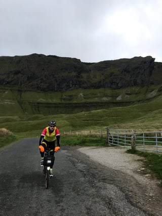 Brompton rider in a yellow gilet rides below a dark cliff-topped mountain on an overcast day heading for Sligo.