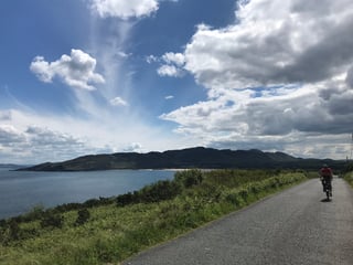 Rider on a loaded Brompton follows a narrow lane beside a calm bay with dark hills behind, west Ireland.