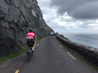 Brompton rider on a narrow cliff road beside a low stone wall above the sea on the Dingle Peninsula.