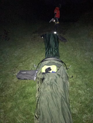 Rider walks in the dark past a bivvy bag pitched on grass beside a TransAtlantic Way Race camp spot.