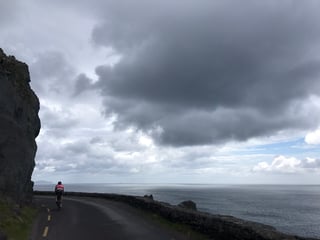 TransAtlantic Way rider on a Brompton cycles a cliffside coastal road below dark rock and heavy cloud in Ireland.