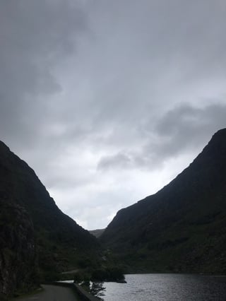 A narrow road runs beside a lake between two steep, dark mountainsides under low grey cloud on the TransAtlantic Way.