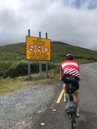 Brompton rider passes a yellow "Turn back now" sign warning large vehicles off a mountain road in Kerry.