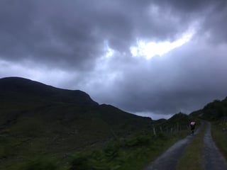 TransAtlantic Way rider climbs a gravel mountain track at dusk beneath dark cloud in Ireland.