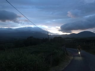 TransAtlantic Way rider with a bike light on climbs a quiet Irish lane at dusk, with hills behind.