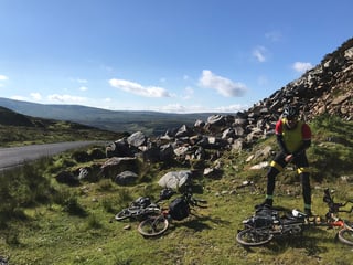 Rider stands by two Bromptons lying on grass beside a rocky roadside bank, wind turbines on the hills behind.