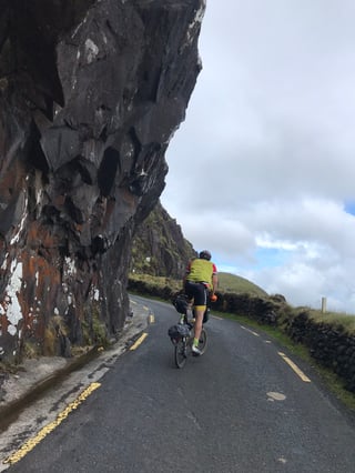 Brompton rider climbs past an overhanging rock face on a narrow road in Kerry, TransAtlantic Way Race.