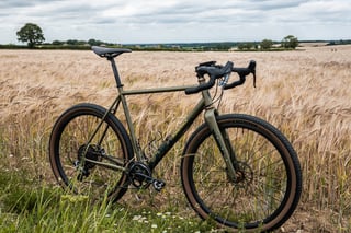 Drive-side view of the Bombtrack Hook EXT in a barley field, with trees and hedgerows on the horizon.