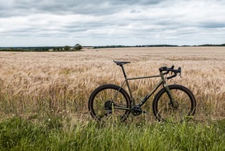 Wide view of the Bombtrack Hook EXT at the edge of a barley field under a grey, cloudy sky.