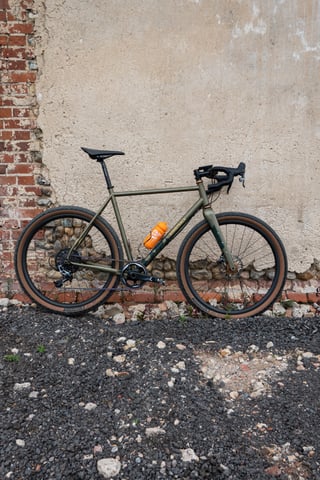 Bombtrack Hook EXT in matte olive leaning against a brick and flint wall, with an orange bottle and tan-wall tyres.