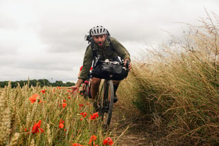 Bikepacker with a handlebar bag rides a field-margin bridleway between wheat and poppies on King Alfred's Way.