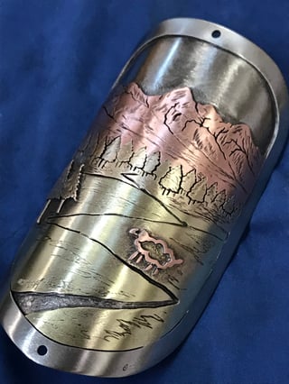 Handmade mixed-metal head badge by Jen Green showing a copper mountain, pine trees, a lake and a small sheep.