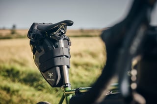 Apidura saddle pack strapped under the saddle of a green Fairlight Faran 2, with a field behind.