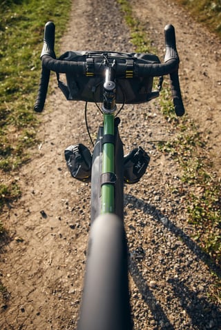 Top-down view along the Faran's green top tube, with Apidura handlebar pack and fork packs on a dirt track.