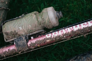 Close-up of a mud-caked bottle cage and Brother Cycles down tube on a gravel bike lying in the grass.