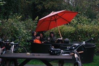 Riders in hooded jackets shelter under a red parasol at a pub garden on the Brother Cycles Kent gravel ride.