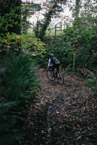 Rider descends a muddy, leaf-covered bridleway past ferns and a waymarker post on the Kent gravel route.
