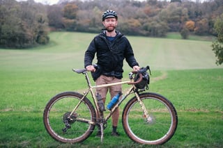 Rider in a mud-spattered jacket with a cream gravel bike and bar bag in a green field edged by autumn woodland in Kent.