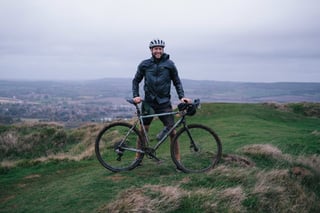 Smiling rider with a muddy grey-green gravel bike on a grassy ridge above the Kent countryside.