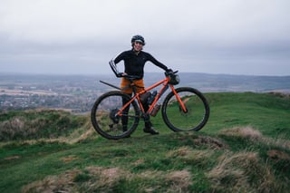 Rider in orange shorts stands with an orange gravel bike and bar bag on a grassy hilltop above the Kent countryside.