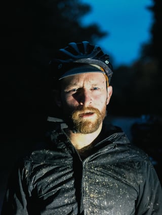 Bearded rider in a muddy black jacket and helmet at dusk, tired after the long Kent gravel ride.