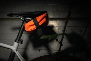 Fluro orange Wiz Viz Teeny Houdini pouch strapped under the saddle of a gravel bike, lit against a dark wall.