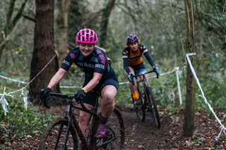Two riders in club kit ride a muddy cyclocross course through woodland, marked out with tape between the trees.