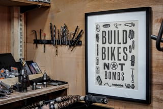 Framed 'Build Bikes Not Bombs' print on the wall of Stayer's workshop, with drill bits and tools beside it.
