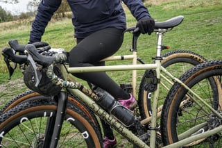 Rider straddles a sand-coloured Stayer gravel bike with camouflage bar tape and a bar bag on grass in Epping Forest.