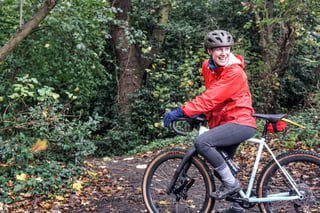 Smiling rider in a red jacket on a pale green gravel bike on a leafy, muddy track in Epping Forest.